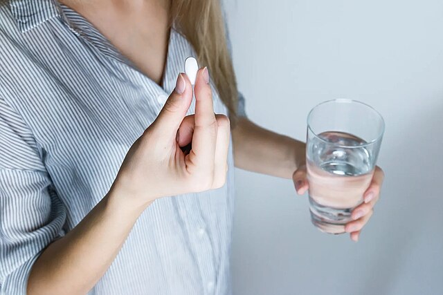 A person holding a white herbal capsule in one hand and a glass of water in the other, preparing to take the medication. The individual is wearing a striped shirt, and the focus is on the hands and the capsule against a plain background.