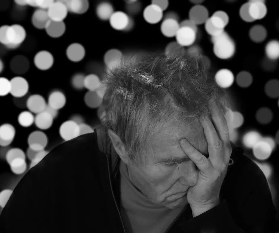 An elderly person with messy hair sitting against a dark background filled with large, out-of-focus light circles, resting their head on one hand in a thoughtful or weary pose.