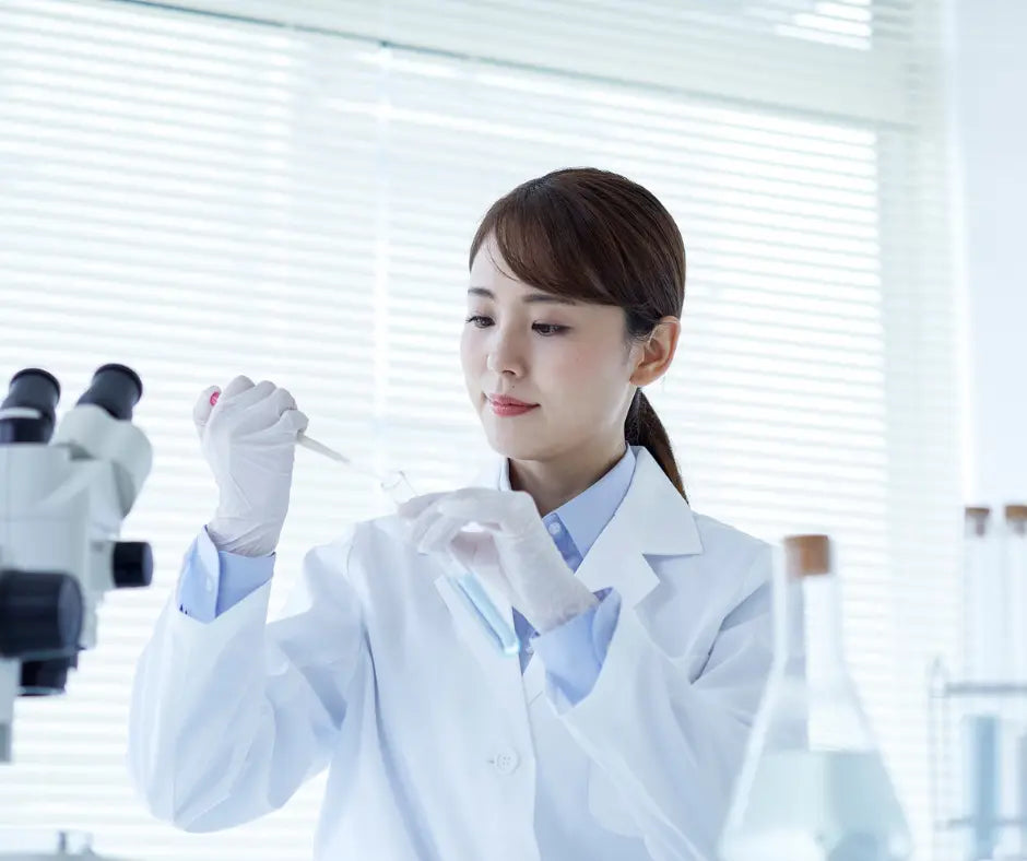 A scientist in a white lab coat and gloves working with a pipette and test tube in a laboratory, laboratory equipment and microscope visible in the background.